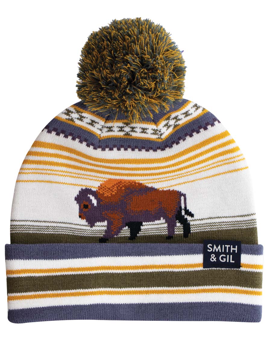 A striped beanie hat with a pom pom on top, featuring a bison, predominantly white, yellow, and brown, with the brand name 'Smith & Gil' on the side.