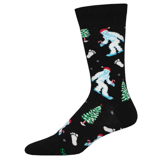 A pair of black crew socks with a design featuring a white yeti wearing a Santa hat, surrounded by Christmas trees and snowflakes.