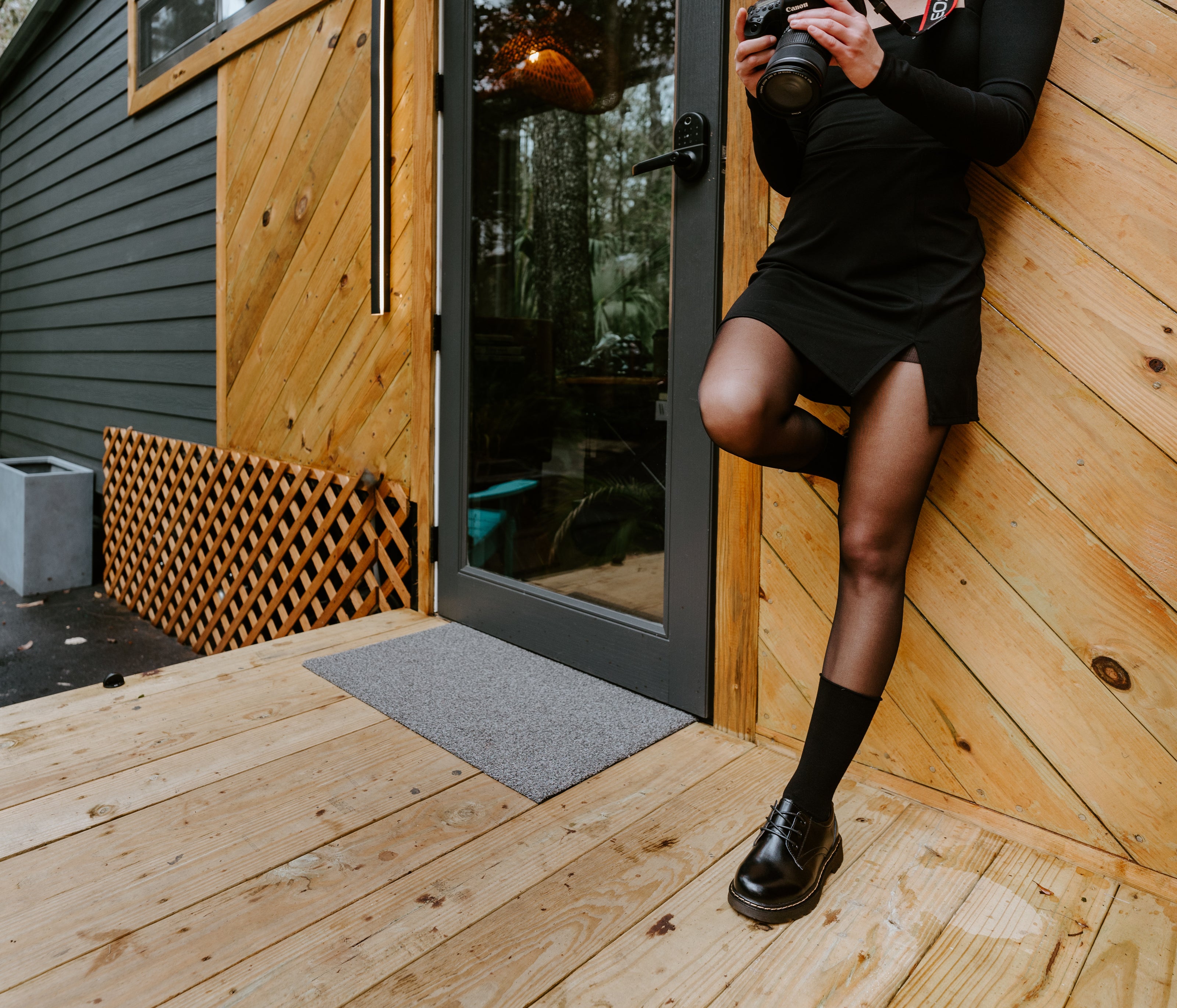 A person in a black long-sleeve mini dress, black tights, black roll-top bamboo socks, and black Mary Jane shoes leans against a wood-paneled wall on a wooden deck, holding a camera.