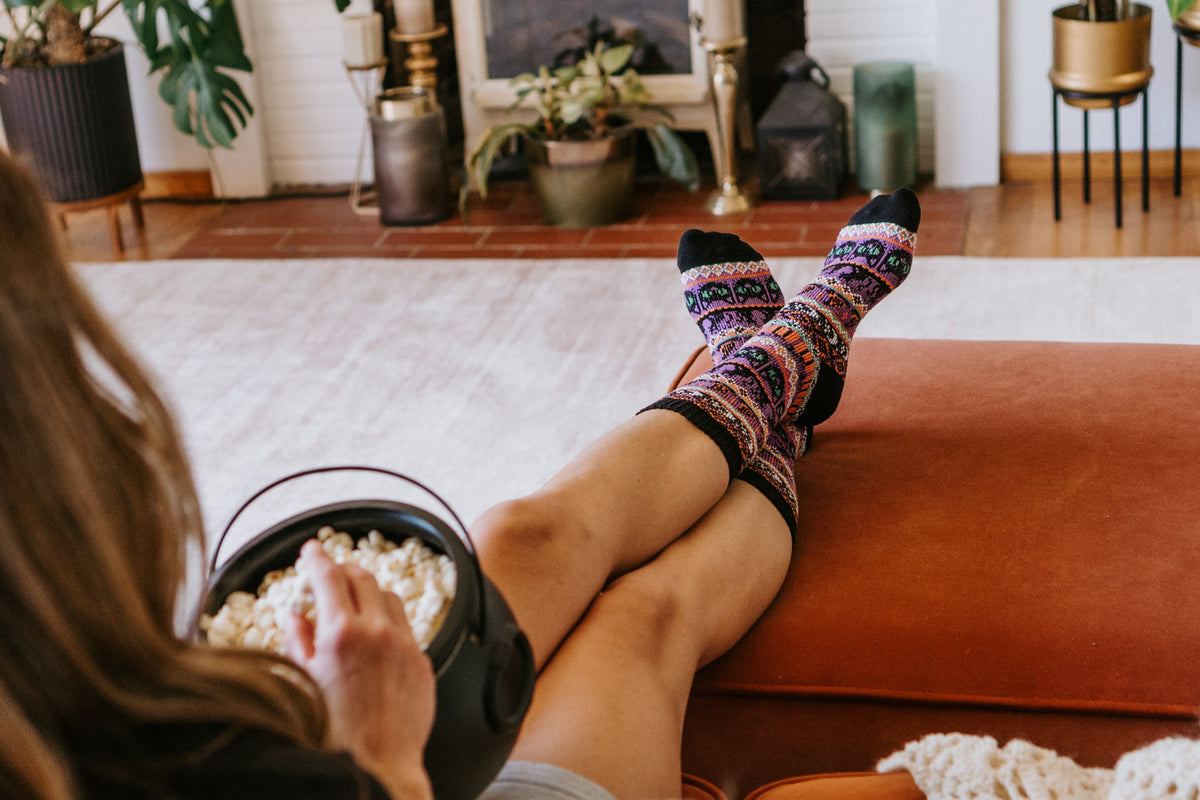 Close-up image of woman wearing Black Cat Voodoo knitted socks in purple and black eating popcorn on sofa