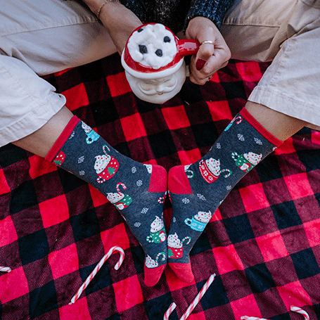 A person wearing festive socks with a mug on a red and black checkered blanket.