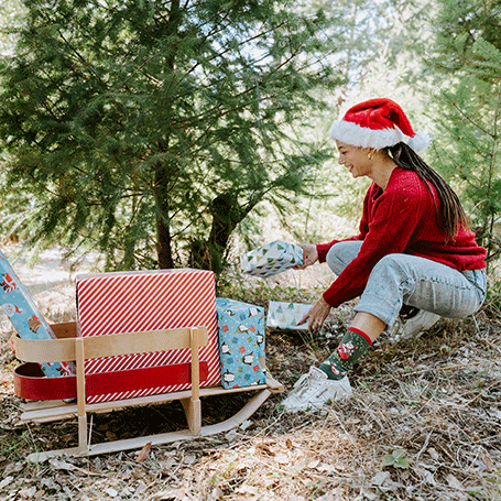 Person in a red sweater and Santa hat sitting in a forest with Christmas presents.