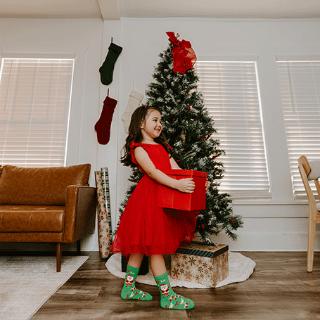 Child in a red dress standing next to a Christmas tree with stockings and presents.