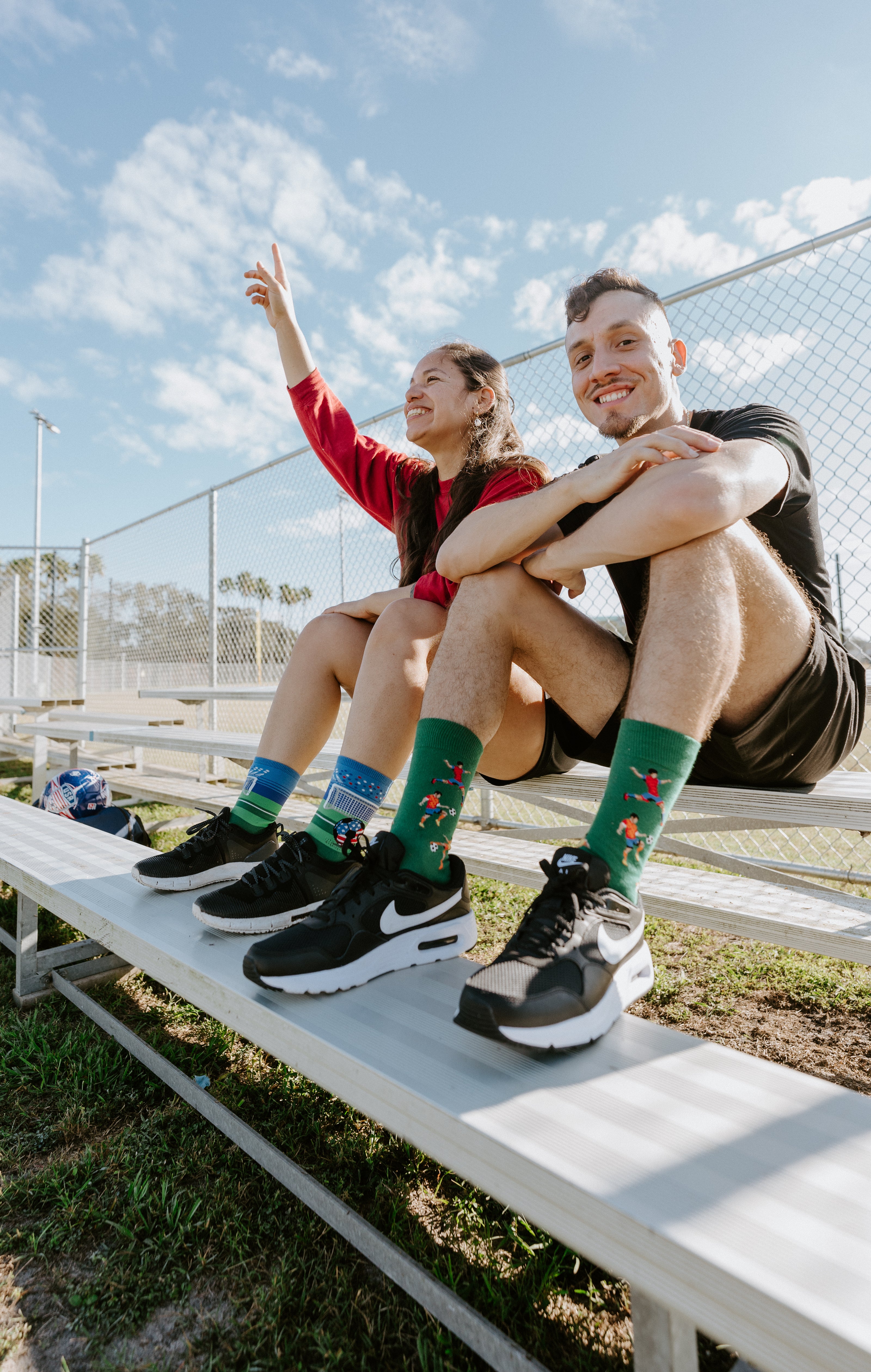 A man sits on bleachers wearing Kicking It socks. Next to him a woman is waving and wearing Soccer's Biggest Stage socks.