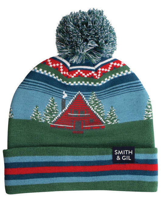 A colorful beanie with a pom pom on top, featuring patterns of trees and an A Frame Cabin.