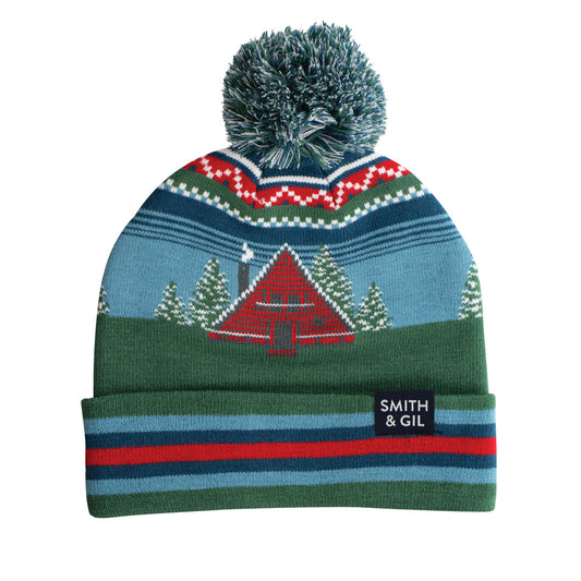 Colorful knit beanie with a design of a cabin and trees, featuring a pom-pom on top, on a white background. Brand 'Smith & Gil' visible.