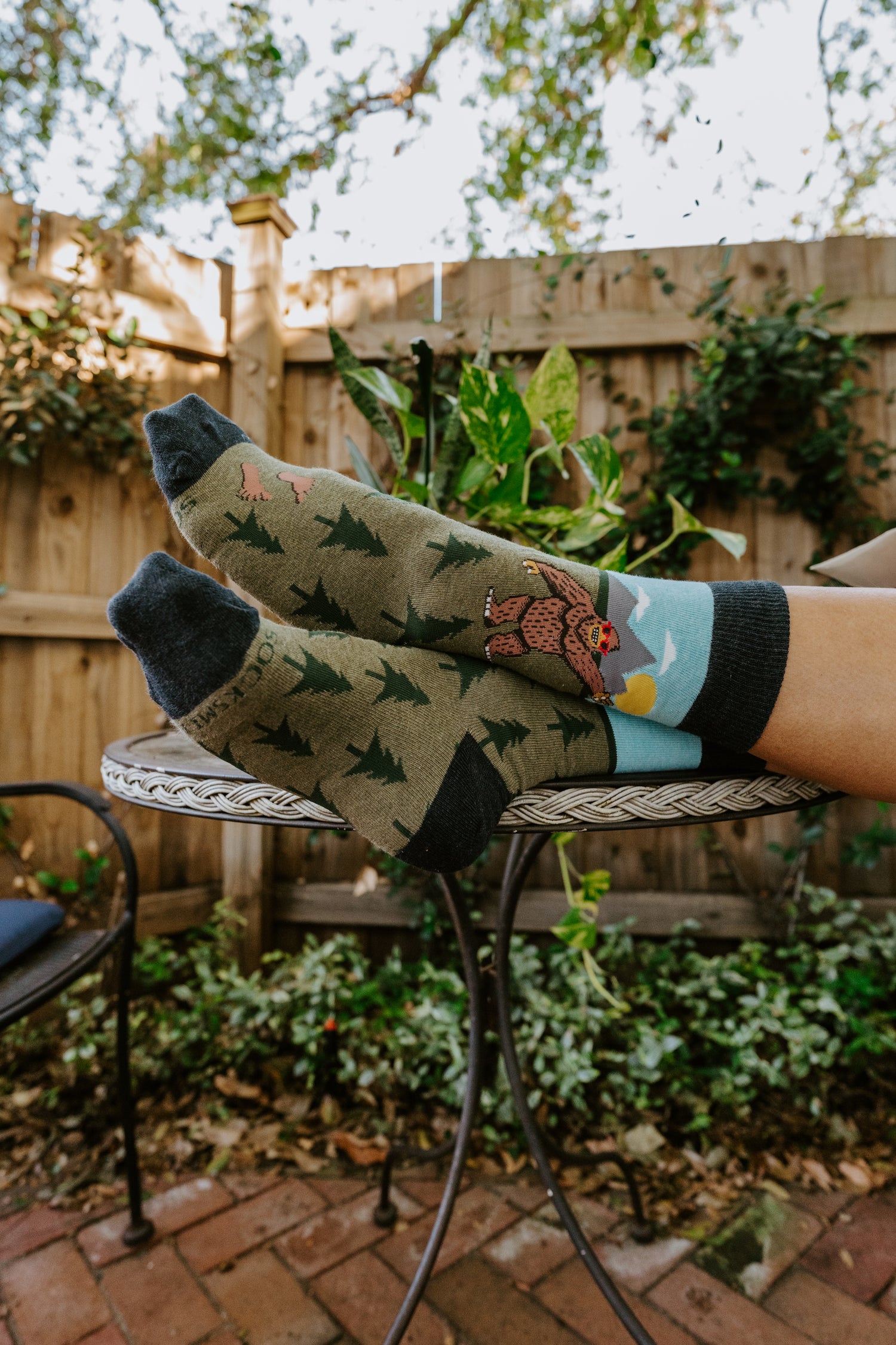 Person wearing colorful socks with nature and bigfoot-themed designs outdoors on a patio.