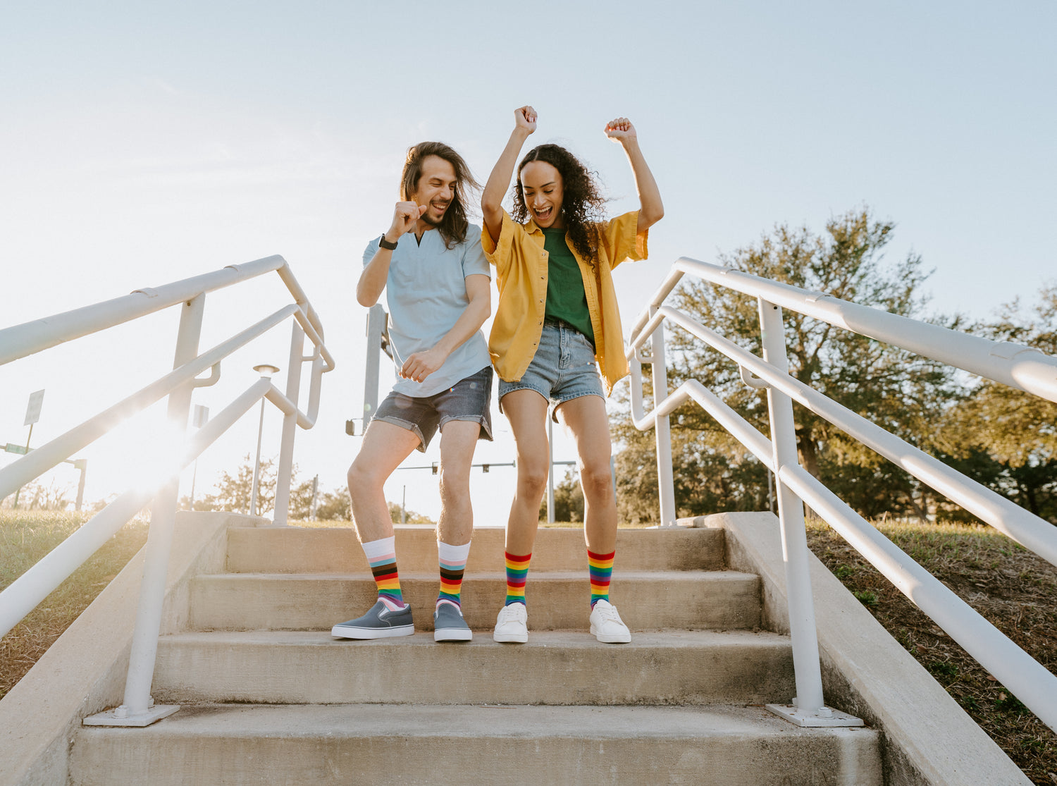 Two people on outdoor steps on a sunny day laughing and bumping hips. The person on the left has long straight hair and a beard, wearing a blue t-shirt, jean shorts, Progress Stripes socks, and blue slide-on shoes. The person on the right has long curly hair, a yellow overshirt and a dark green t-shirt, jean shorts, Pride Stripes socks, and white sneakers.