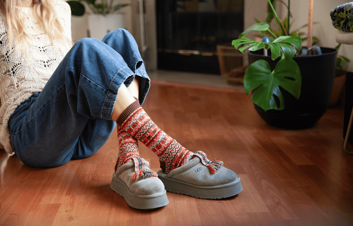 Close-up image of woman wearing seasonal Sweater Socks in Pumpkin Spice Is Nice theme while sitting by fire and plant