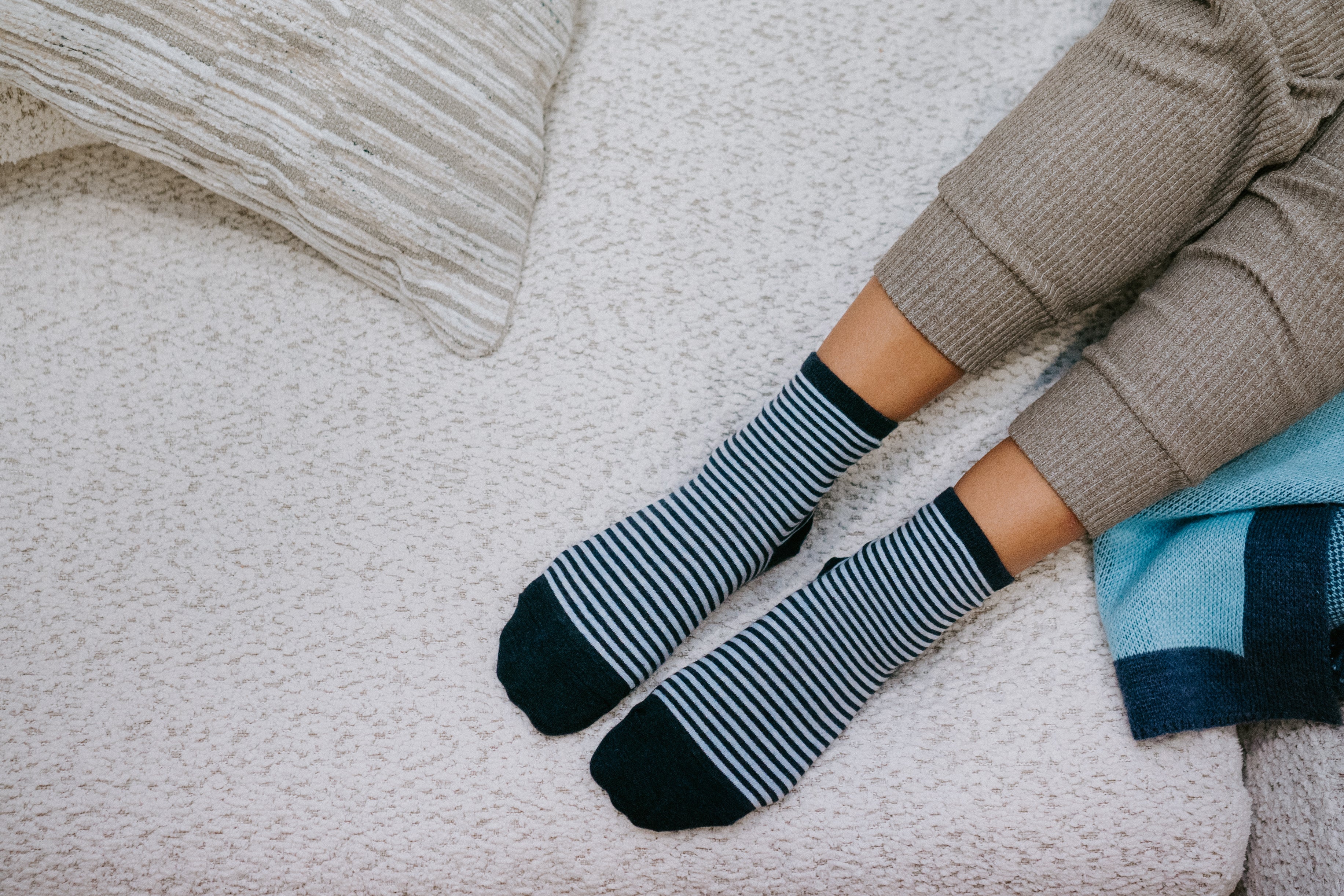 A person wearing striped crew socks with solid navy toes and heels, sitting on a textured cream blanket next to a pillow and a blue throw blanket.