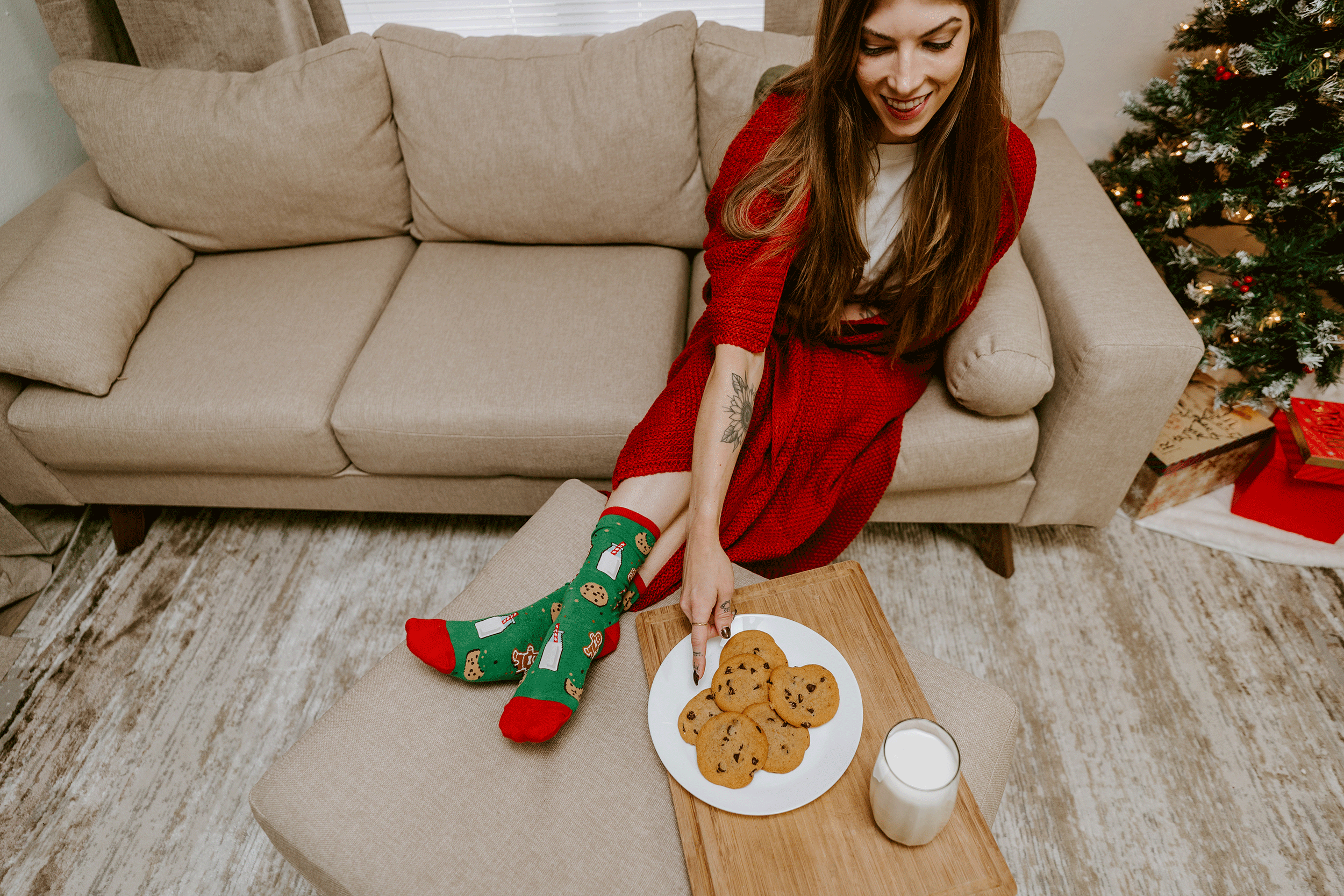 Woman in a red dress sitting on a couch with cookies and a glass of milk, Christmas tree in the background wearing Christmas Milk and Cookies themed socks