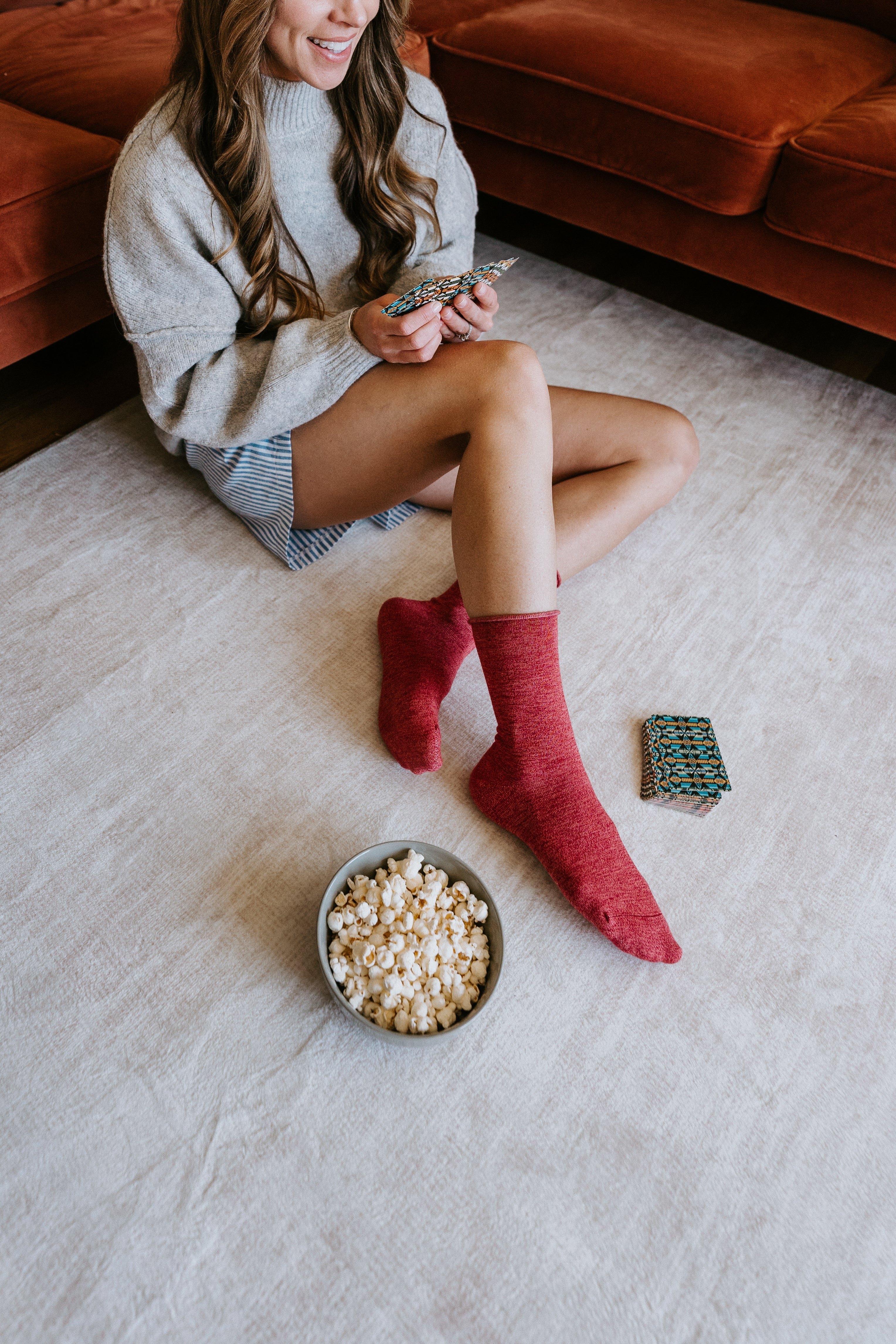 A woman sits on a light beige rug in a living room, wearing a chunky light gray knit sweater and red super-soft bamboo socks while holding cards. A bowl of popcorn and a deck of cards are on the floor in front of her, with a rust-colored sofa in the background.