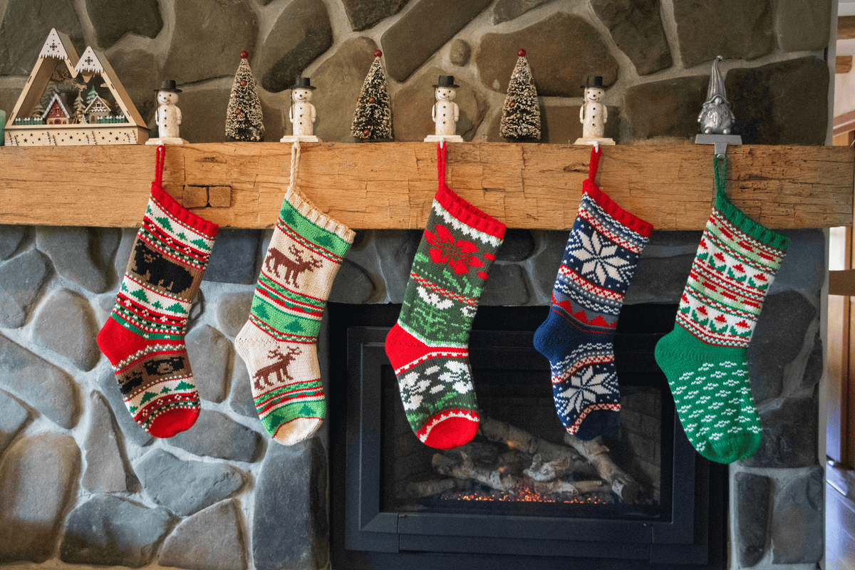 Five festive Christmas stockings with colorful knit patterns hang from a rustic wooden mantel above a stone fireplace. The stockings feature holiday motifs including snowflakes, reindeer, and snow globes, with red, green, blue, and cream colors.