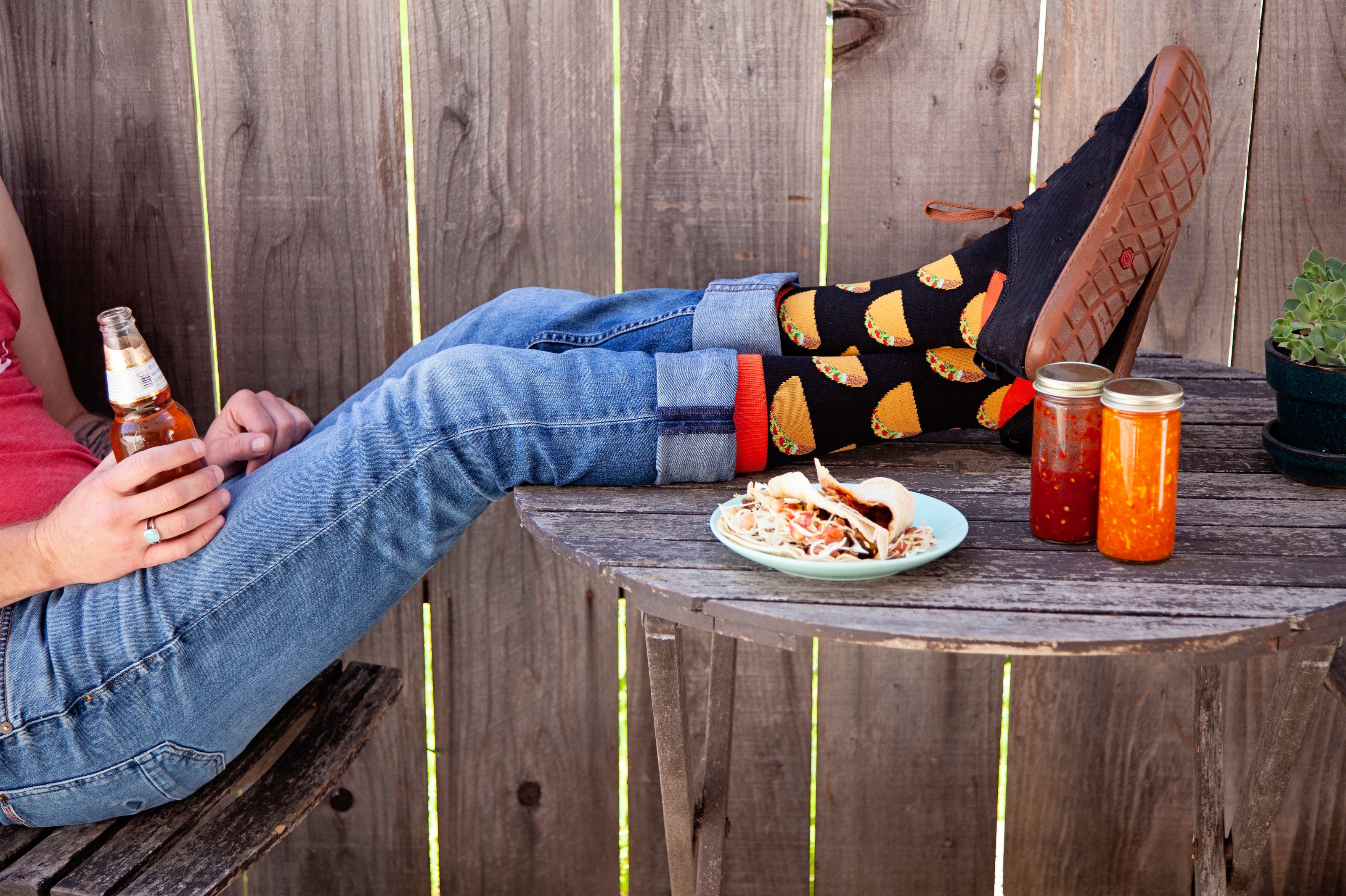 Person sitting on a wooden bench with a beer, food, and drinks on a table wearing Tacos socks.