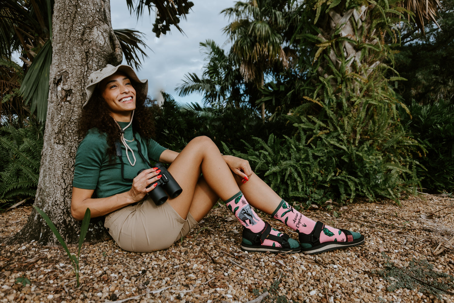 A dark haired woman sitting in front of lush tropical plants and smiling while wearing an outdoor hat and holding binoculars. She's wearing pink Asian Elephant socks and sandals, a green shirt, and tan shorts.