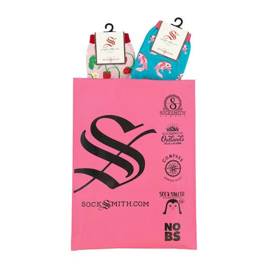 A pink Socksmith gift bag with black printed logos and text, holding two packaged pairs of women's cotton novelty socks.