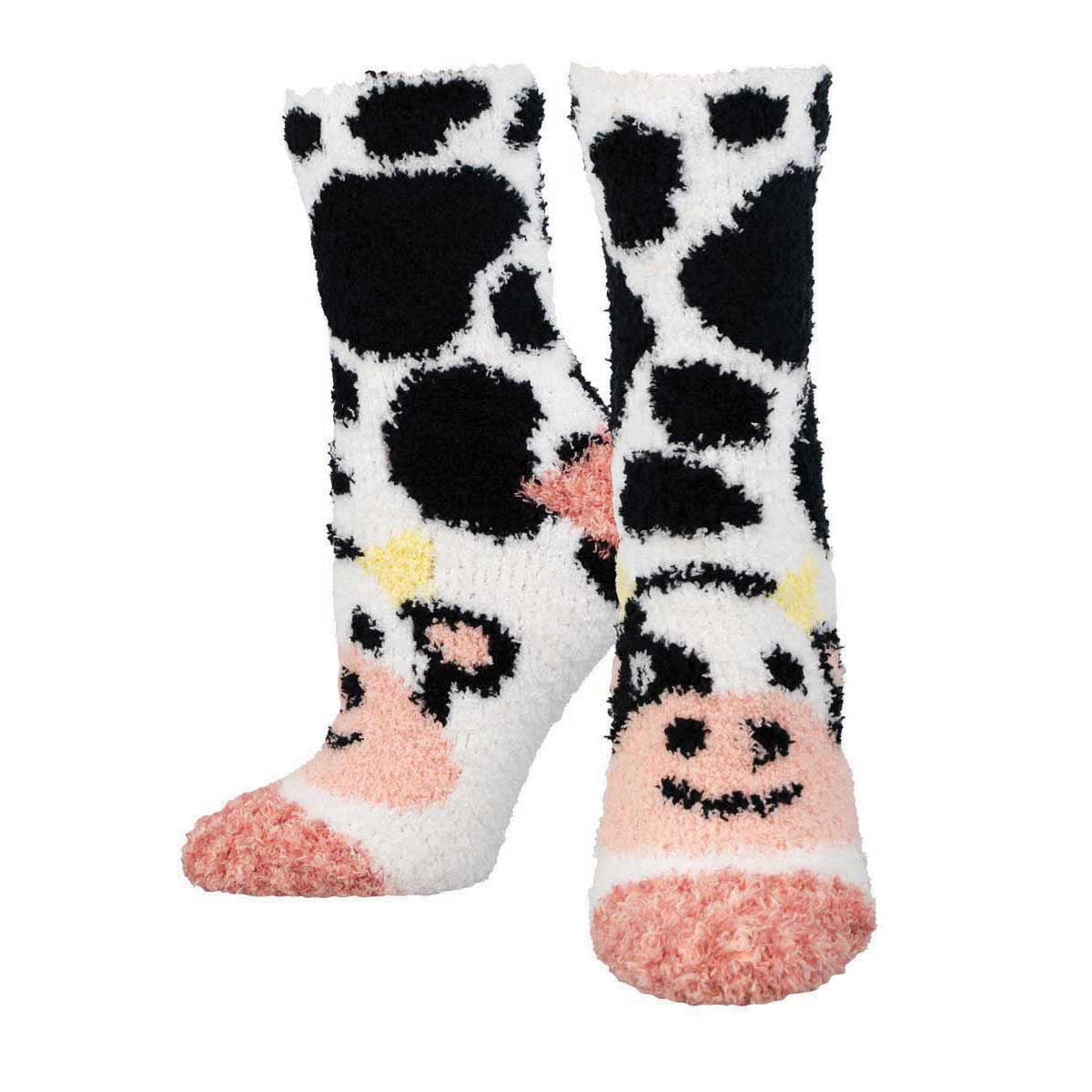 A pair of fuzzy white crew socks with black cow print and a cartoonish cow face design on the heel area, complete with pink accents.